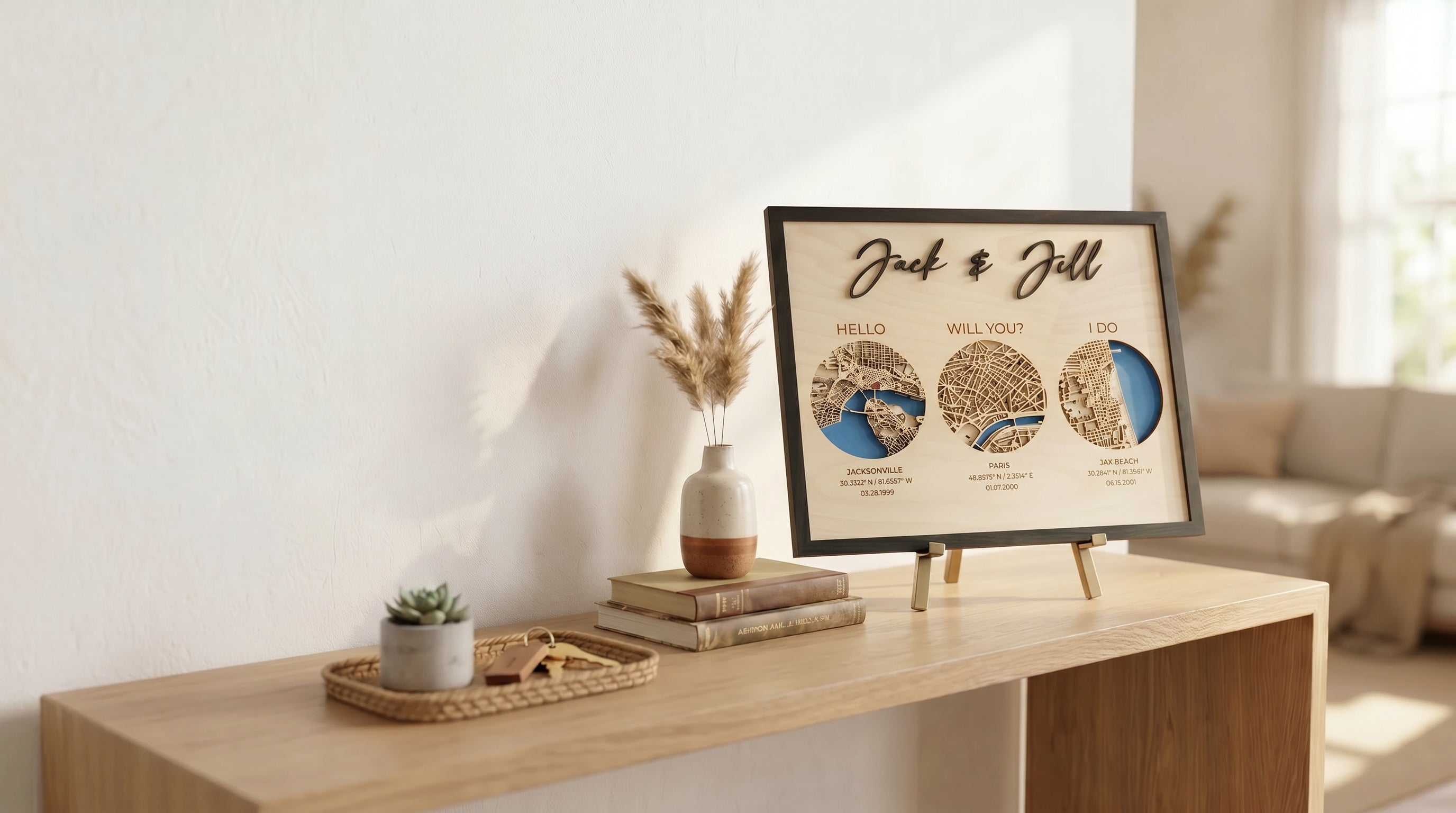 Custom laser-cut wooden wall art featuring a cursive "Jack & Jill" name sign with three circular layered map medallions in blue and natural wood tones. Each circle is labeled with a milestone phrase — "Hello" over Jacksonville FL (03.28.1999), "Will You?" over Paris France (01.07.2000), and "I Do" over Jax Beach FL (06.15.2001) — with coordinates beneath each city. The framed piece sits on a gold easel on a light oak console table styled with a ceramic vase of pampas grass, stacked books, a succulent, and w